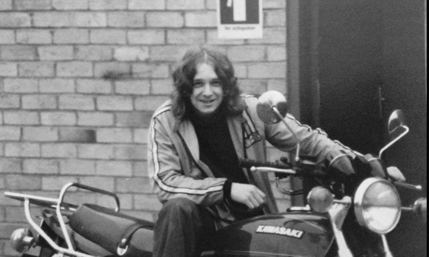 A black and white photograph of a person sitting on a Kawasaki motorcycle in front of a brick wall.